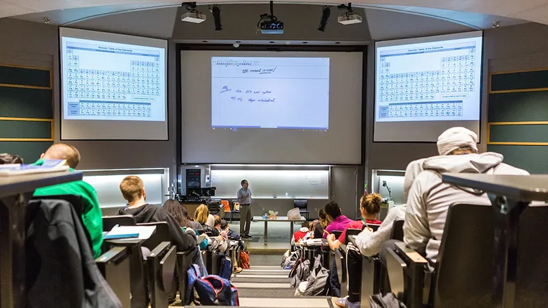 A professor leads a class in a lecture hall, with technology displaying the periodic table and notes.